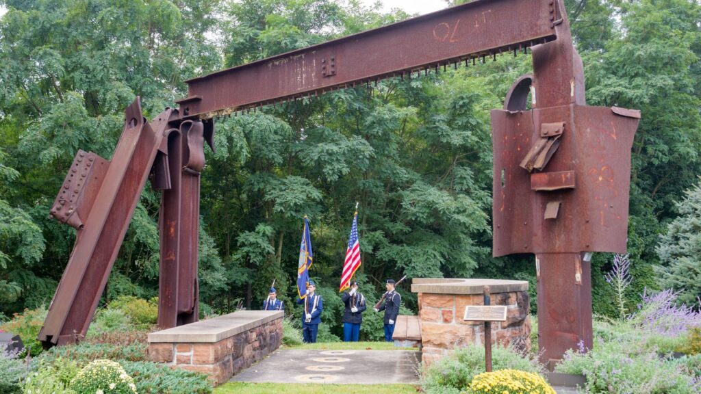 rectangular arch of twisted steel beams to commemorate 9/11 with ROTC cadets in dress blues carrying flags and rifles in the background.