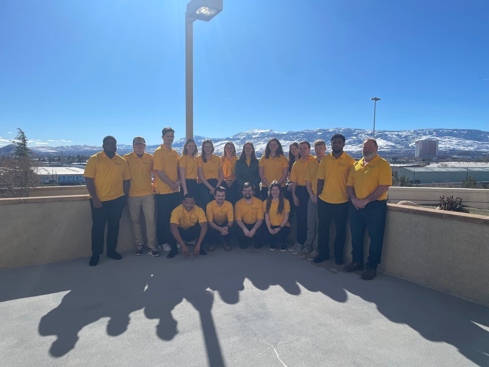 A professor and 16 students wearing yellow polo shirts pose outdoors on the roof of a building with a mountainous background.