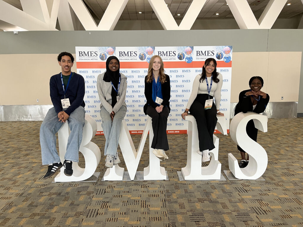 Five students sit or lean on large letters BMES in front of a media backdrop.