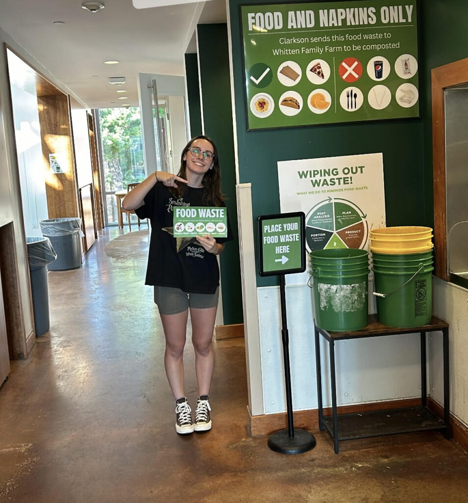 A female student holding a sign that reads "Food Waste" and pointing to green and yellow buckets next to a "Place Your Food Waste Here" sign. A poster reading "Food and Napkins Only, Clarkson sends this food waste to Whitten Family Farm to be composted" is located above the buckets.