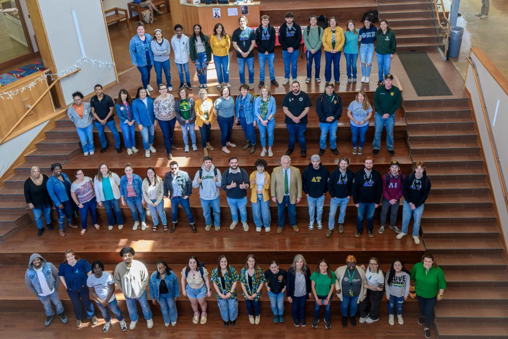 Several dozen people wearing denim are looking up to pose for a photo from the steps of Clarkson's student center