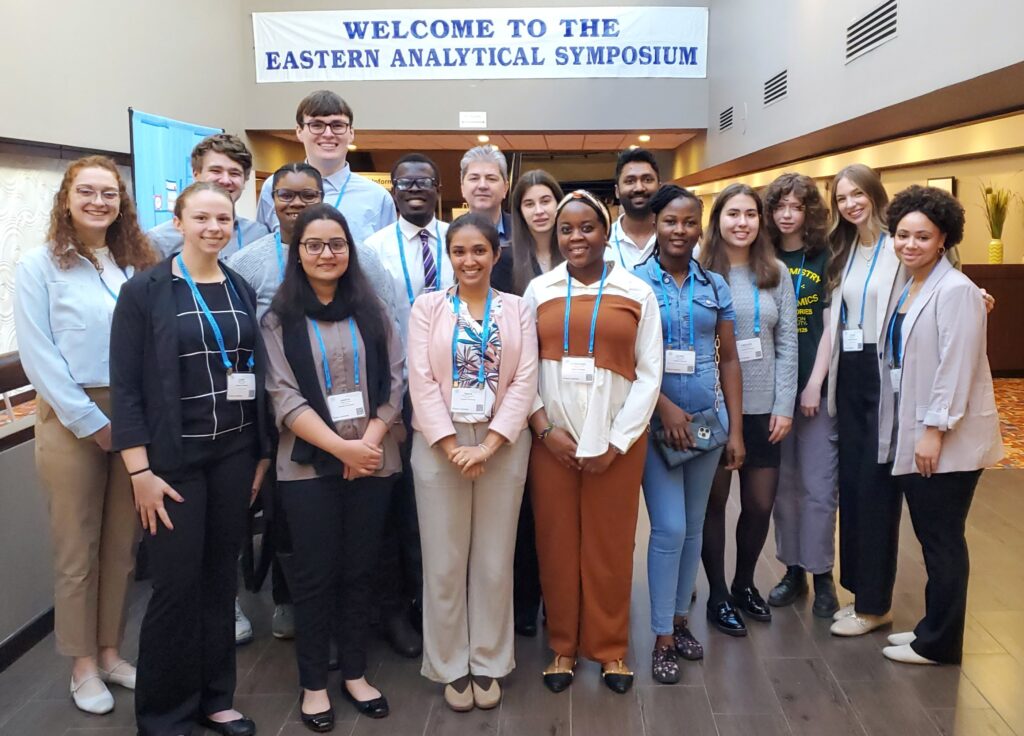 About 16 students pose for a photo with their professor, in front of a banner reading "welcome to the Eastern Analytical Symposium"