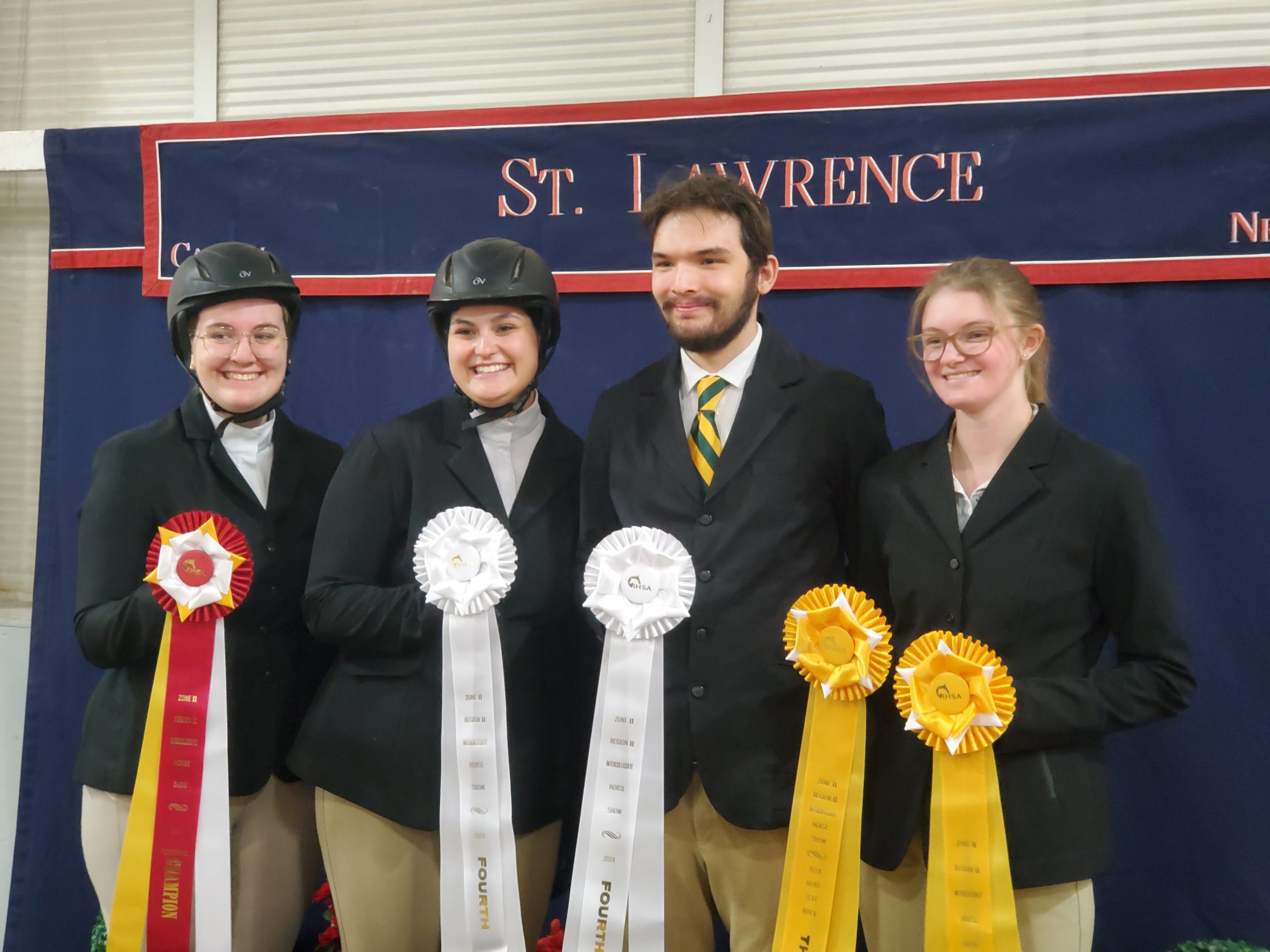 4 people smiling wearing equestrian riding uniforms. Pictured from left to right, a woman holding a second place ribbon, a woman holding a fourth place ribbon, a man holding a fourth place ribbon and a woman holding two third place ribbons.