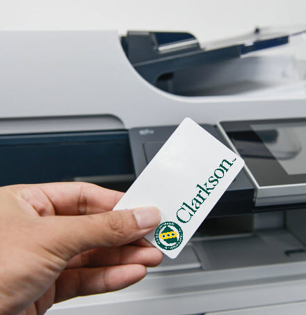 Image of a hand holding a Clarkson ID  card with a printer in the background. Indicates an individual will scan their card at the printer to access to their print job.
