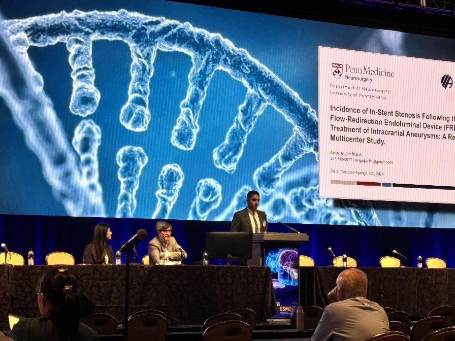 A man in a suit stands on stage with DNA on a projector screen behind him.