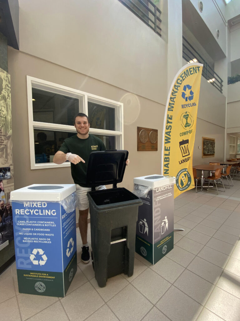 A young man standing next to a mixed recycling bin, a compost toter, and a landfill garbage bin. A flag behind him reads "Sustainable Waste Management."