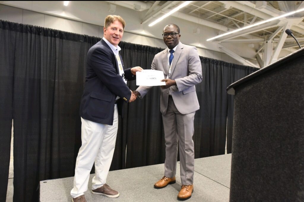 A student shakes hands with a conference organizer on stage as a certificate is held between them.