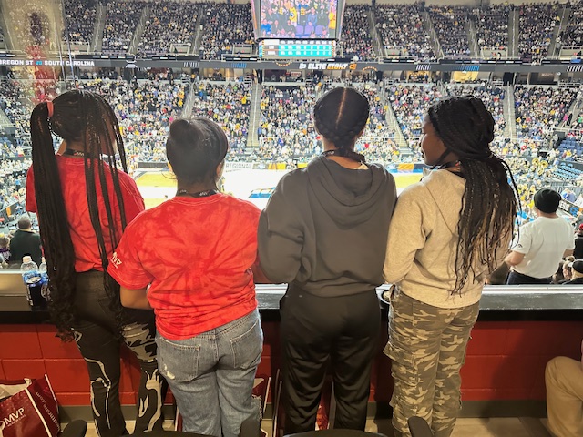 Four girls watch a basketball game in a crowded arena
