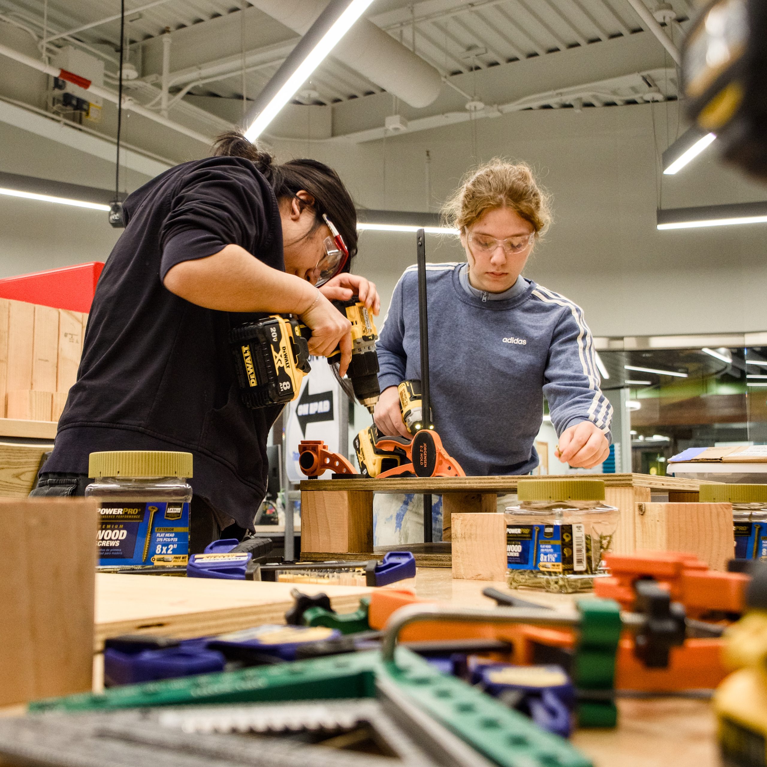 Two Maker Mentors construct a wooden base using drills, rulers, and other woodworking tools.