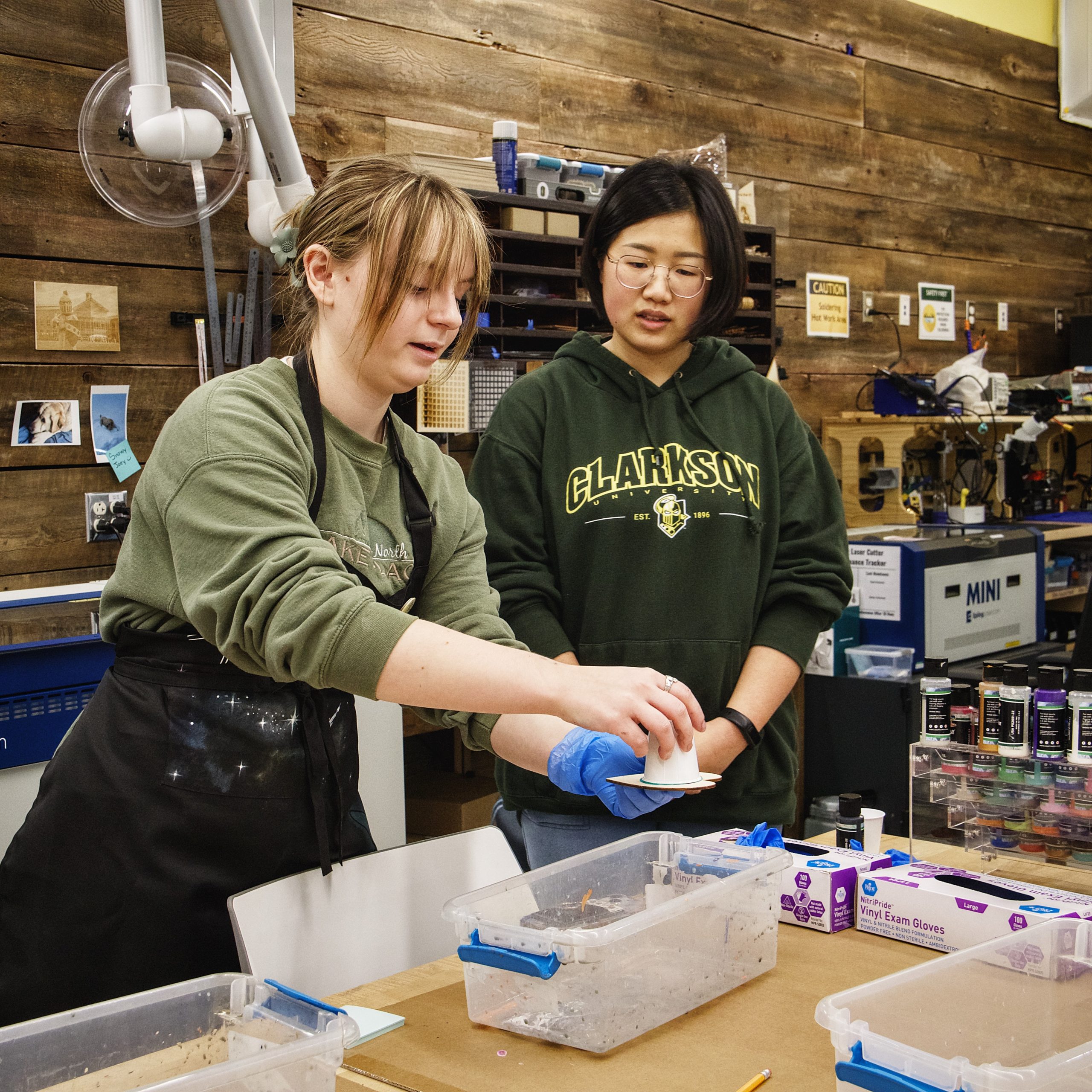 Two students stand in the Makerspace, holding a small paper cup used alongside acrylic paint while creating glass art.