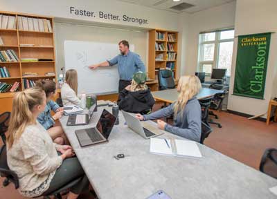 three students sit at a table with their laptops open as a professor points to a white board depicting a line graph with upward projections.