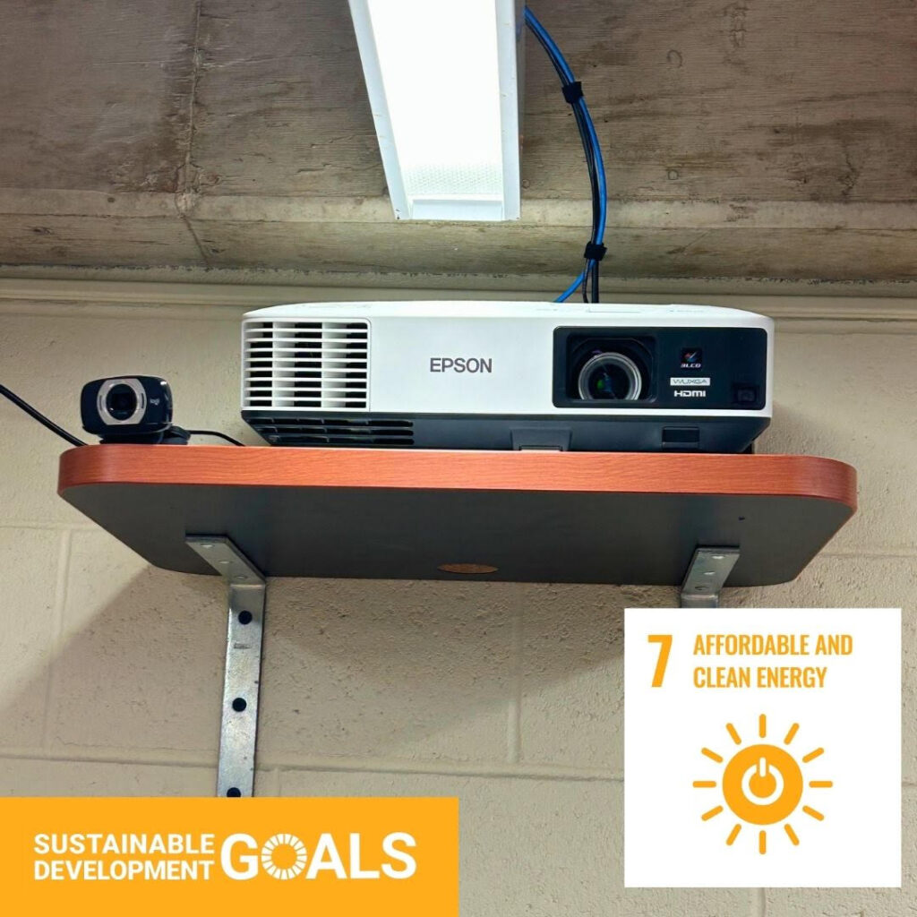 An Epson Projector resting on a wooden shelf, with a small camera next to it. The United Nations "Sustainable Development Goals" logo is pasted on top of the image in an orange rectangle. A white square with the text "7: Affordable and Clean Energy" is featured in the bottom right corner.