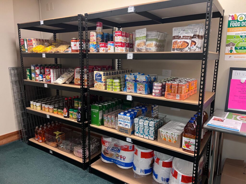 Two shelving units filled with various foods including canned meats, canned fruits, pasta, and sauce as well as hygiene products such as toothbrushes, tampons, and pads.