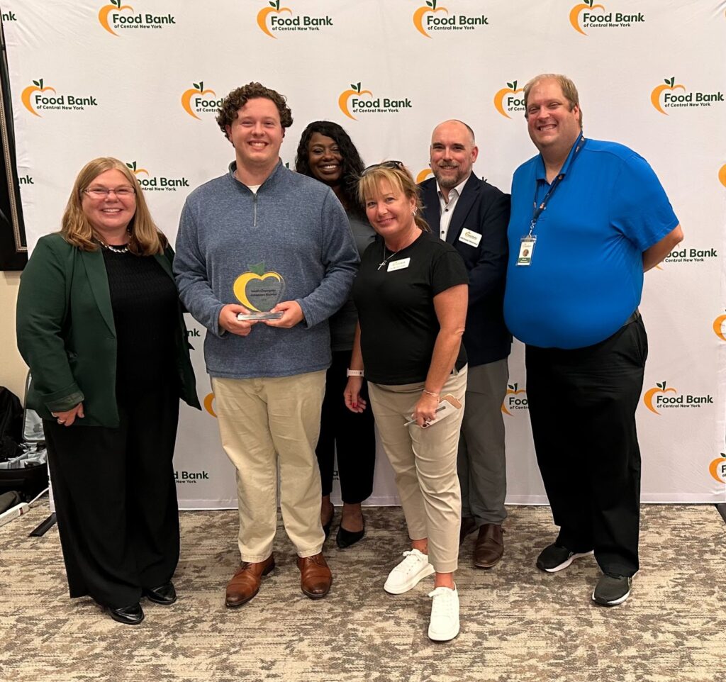 A group of six people stand in front of a Food Bank of Central New York backdrop. One person in the center holds a transparent award shaped like an apple, a nod to Food Bank of Central New York’s logo.