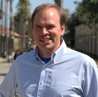 Headshot of Dr. Matthew Wheeler in a blue shirt, and outdoor background