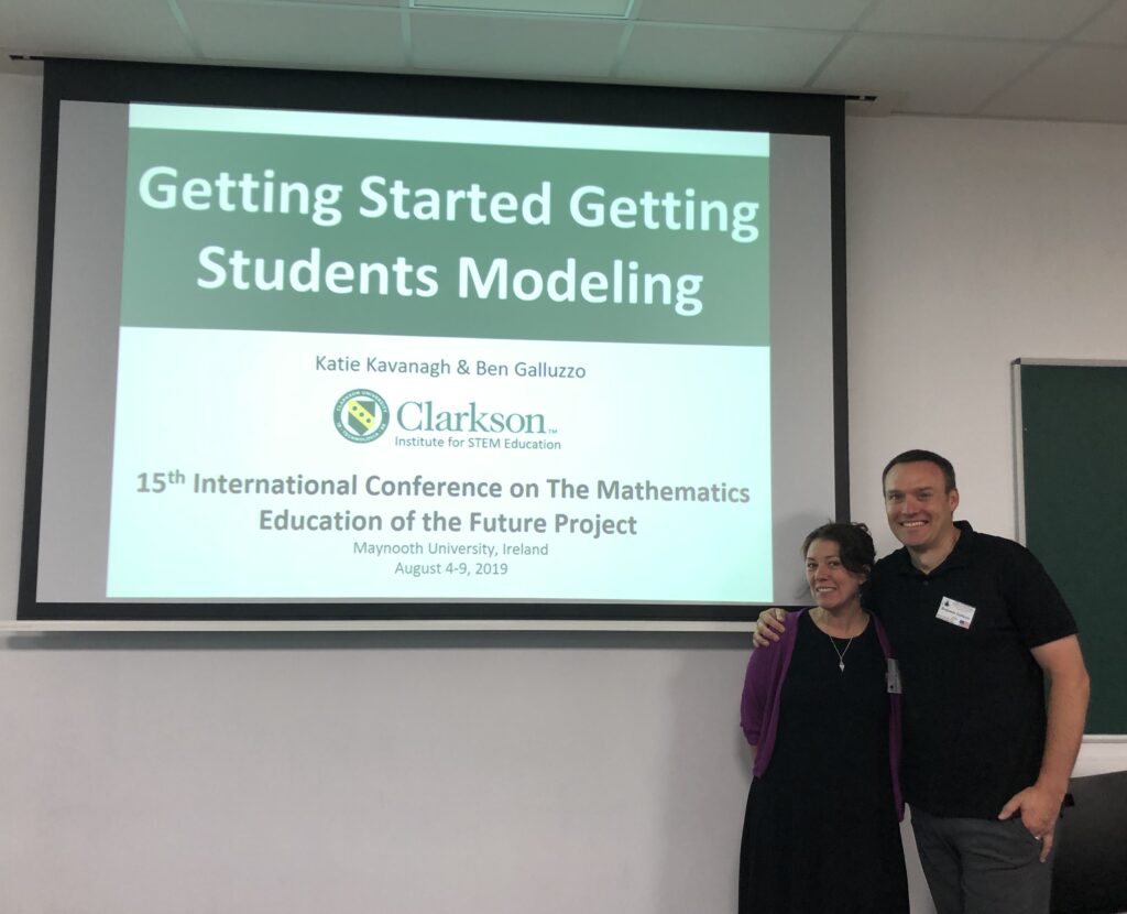 A man and woman pose in a friendly embrace next to a projector screen reading "Getting Starting Getting Students Modeling"