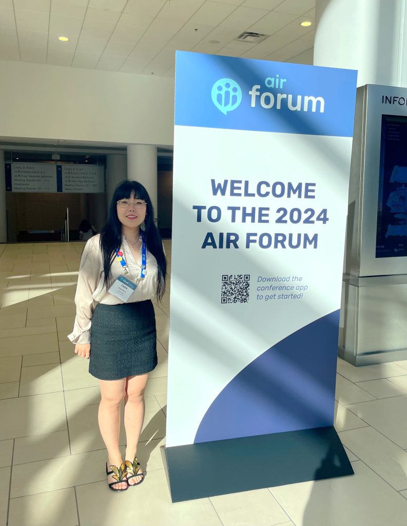 A young woman poses next to a vertical welcome banner for the AIR Forum.