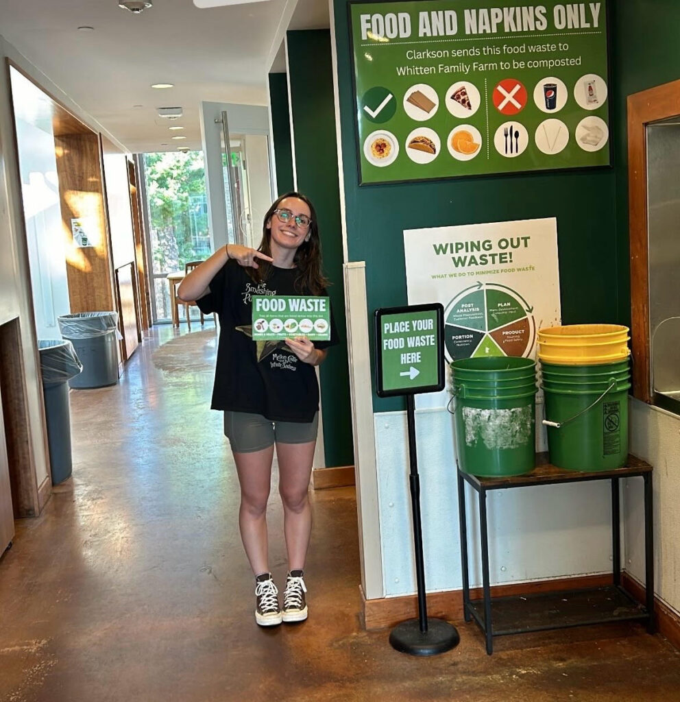 A student standing next to the food waste buckets by the Servery dishwashing window. She is holding a sign that reads "food waste" and is pointing to the buckets and a sign that reads "place your food waste here." There is also a poster behind the buckets indicating what can and cannot be placed in the compost buckets.