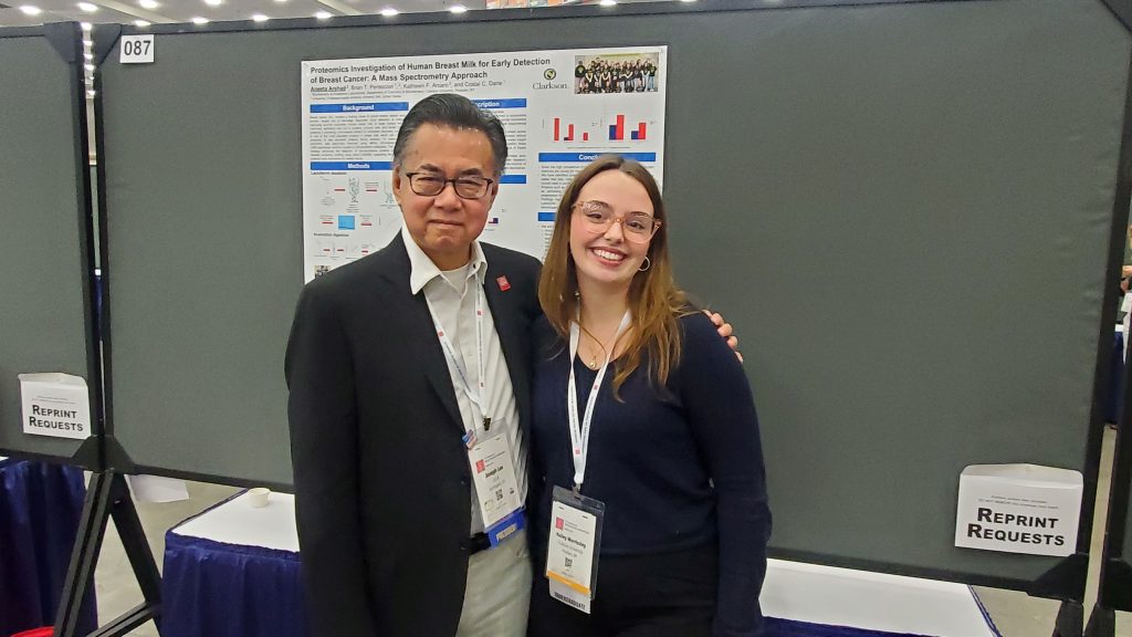 A student stands with a professor in front of a poster presentation as they both pose for a photo
