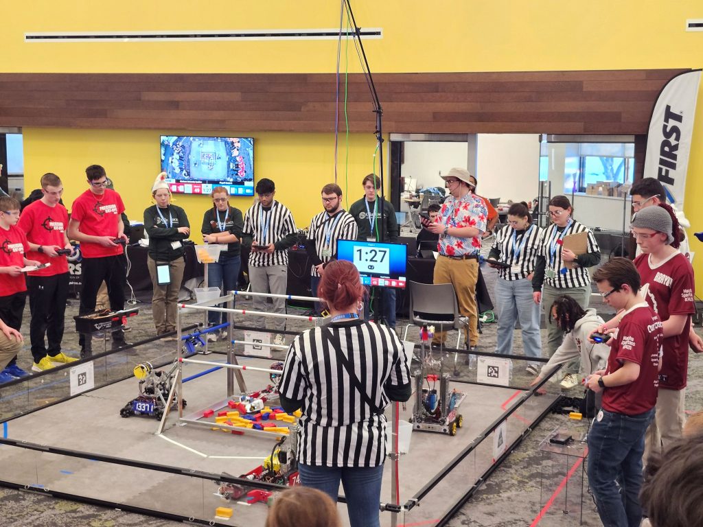 Several students in team t-shirts and referees gather around a robotics ring.