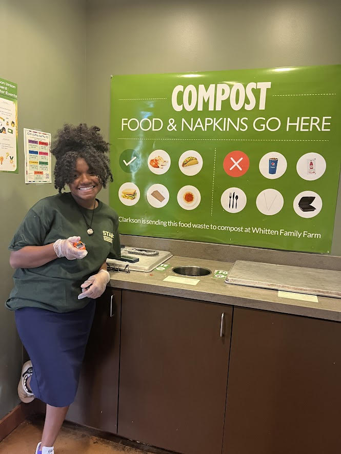 A student standing by the Cafeteria waste bins station, and beside her a poster displaying "Compost; Food and Napkins go here, Clarkson is sending this food waste to compost at Whitten Family Farm"