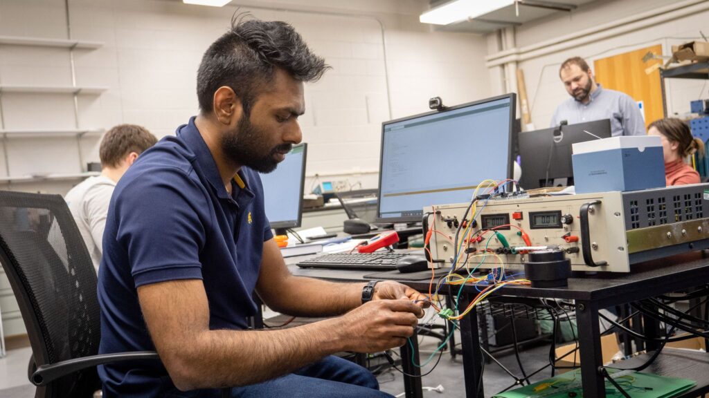 A student sits at a computer connecting wires to a computer hardware box in a lab.