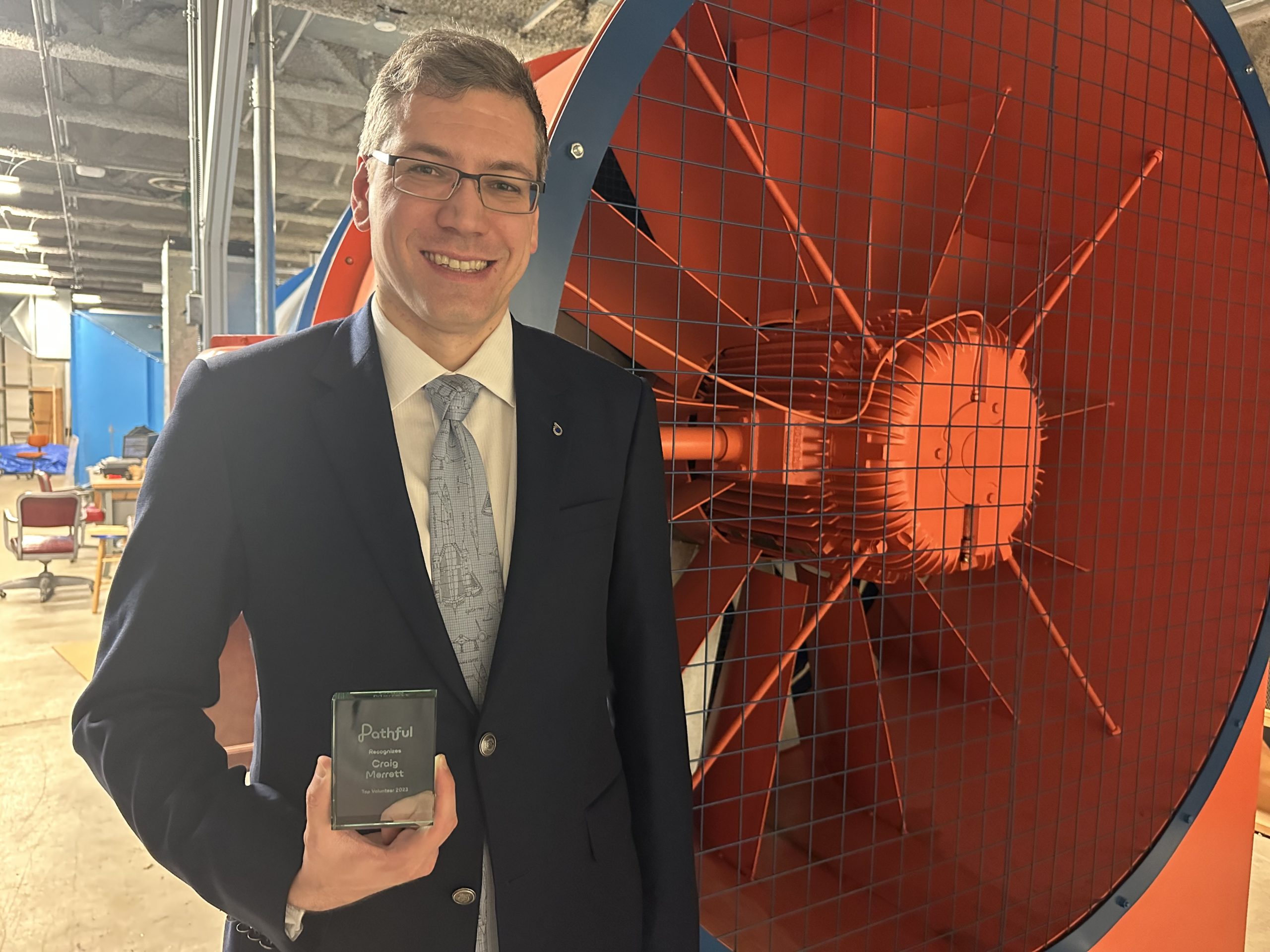 Portrait of Craig Merrett holding a crystal plaque next to a large fan connected to a wind tunnel.