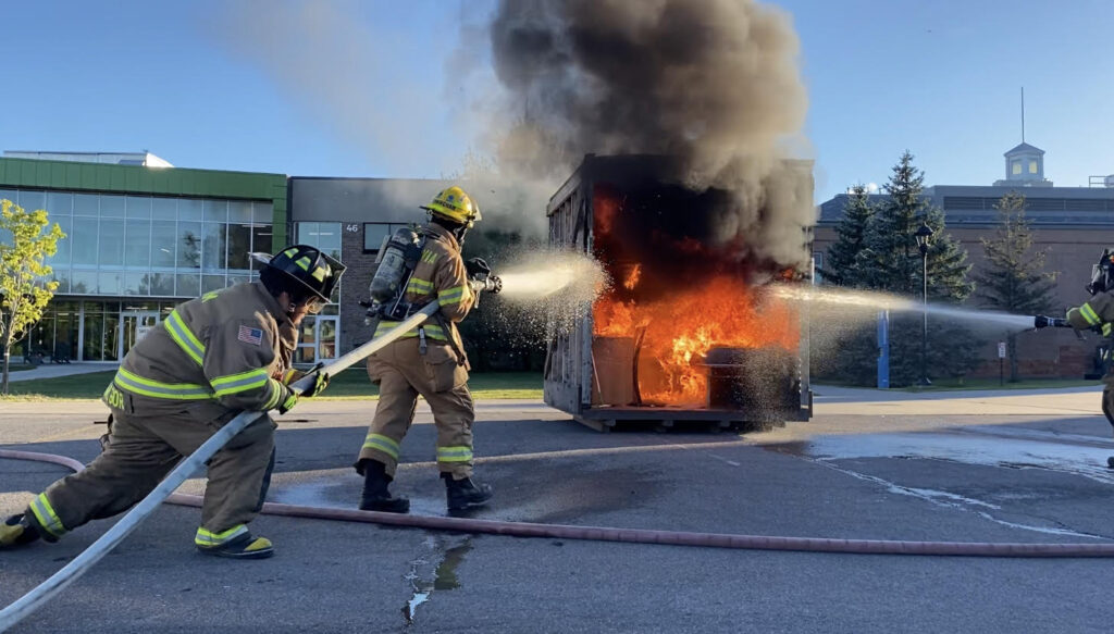 Firefighters spray a fire-filled model dorm room with fire hoses in a parking lot in front of a building on Clarkson's campus.