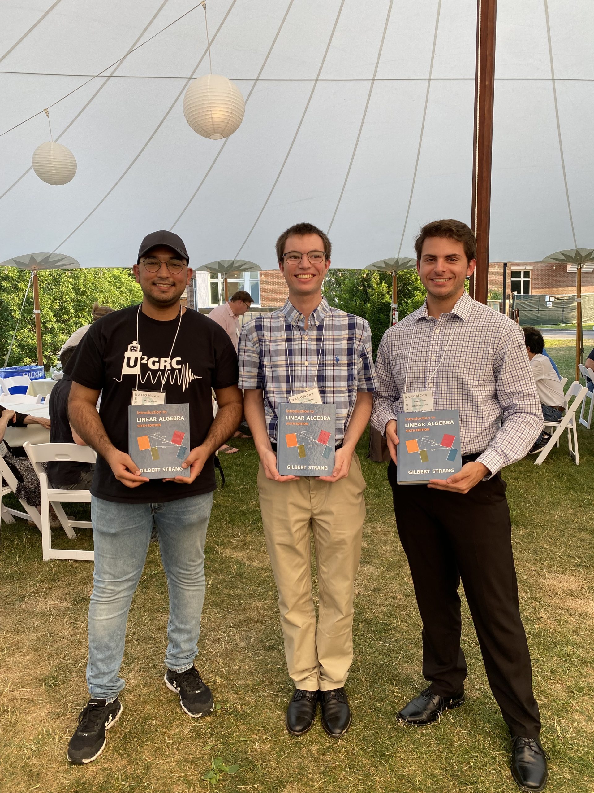 Three men pose with copies of a linear algebra book under an outdoor tent.