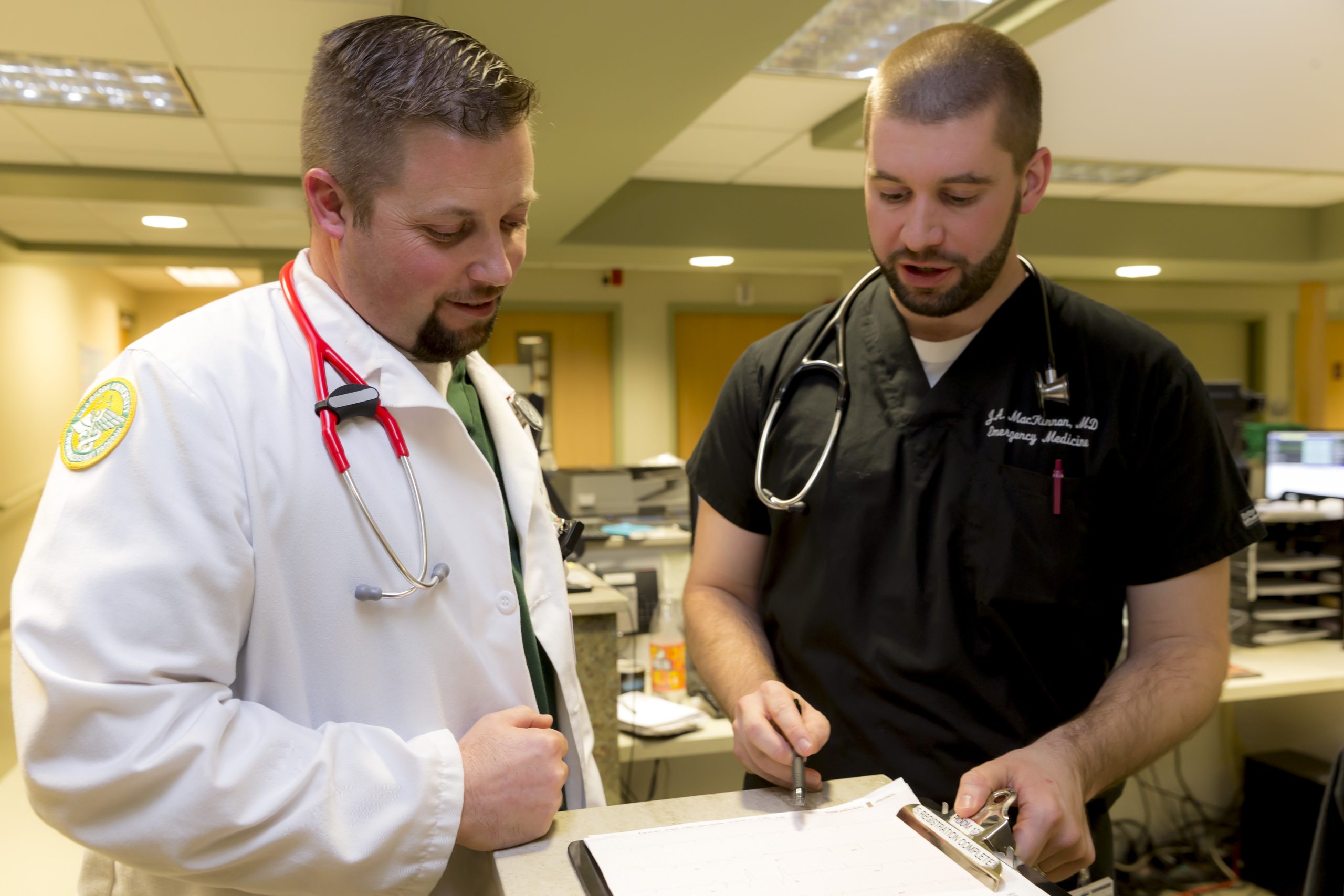 two men in medical garb review a paper on a clipboard.