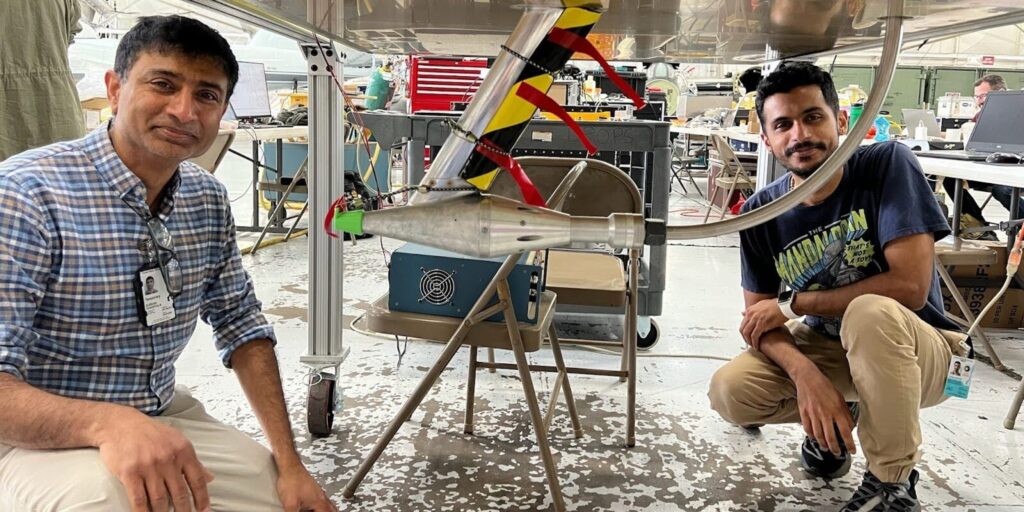 Two men crouch beneath a metal platform from which a particle sampling device is attached.
