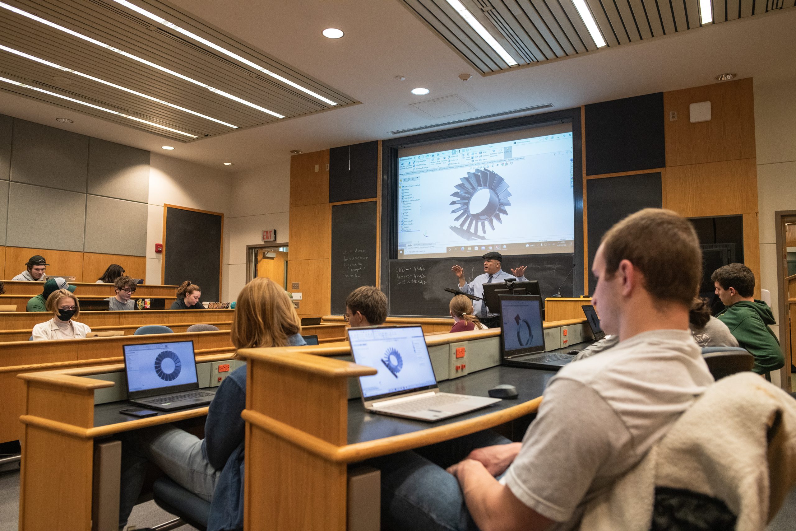 A professor stands at the front of a classroom in front of a projector showing a concept of a gear or turbine. Students sit facing away in the foreground, with the same image displayed on laptops.