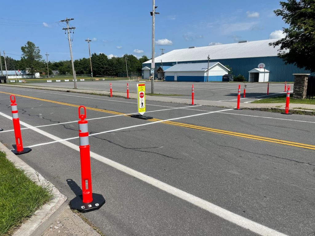 Traffic cones line the sides of the street while a pedestrian crossing sign sits in the middle of the street in a crosswalk with a community hockey rink in the background.
