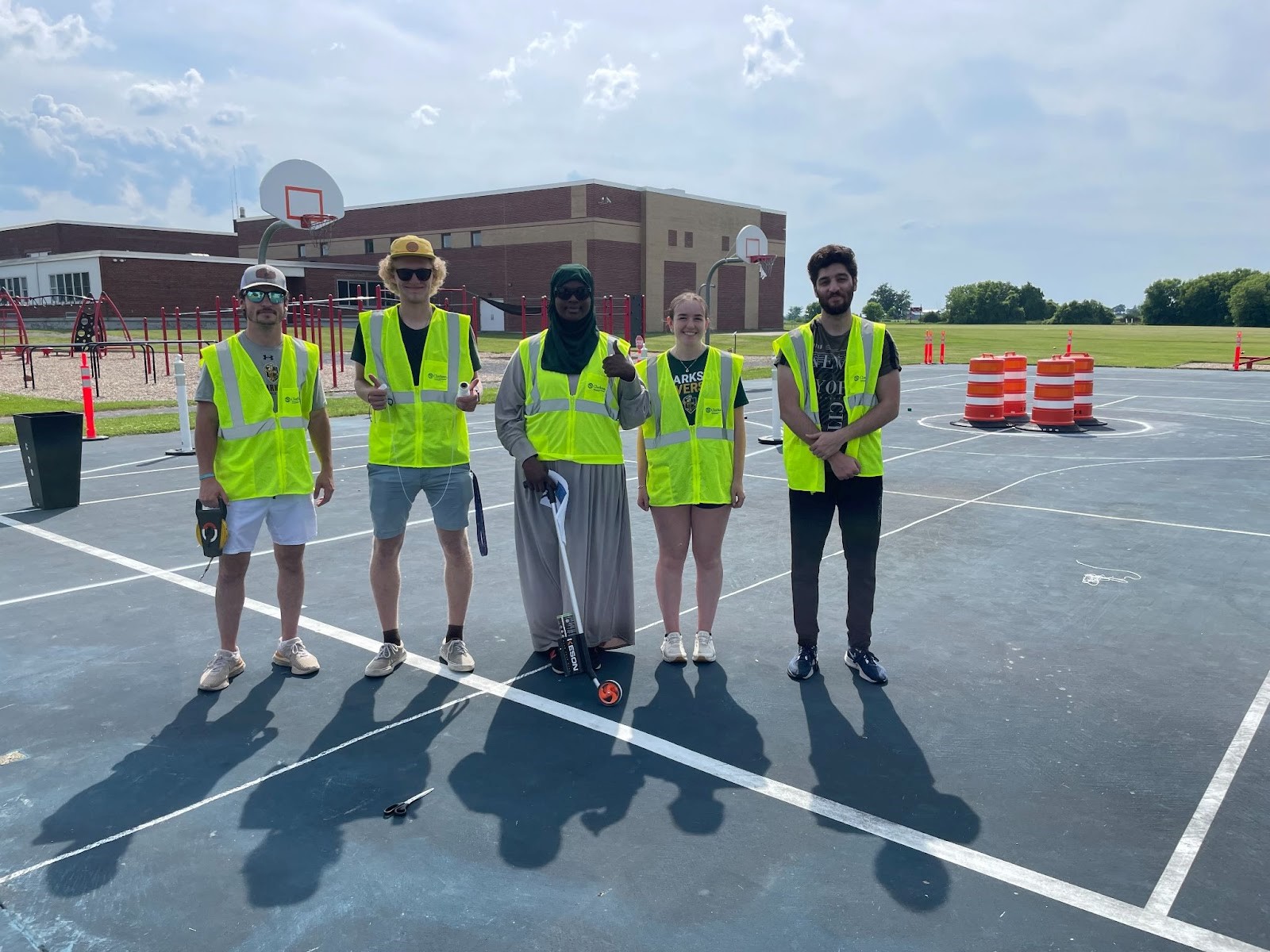 Five students pose for a photo in high-visibility vests on an outdoor basketball court with various traffic cones in the background