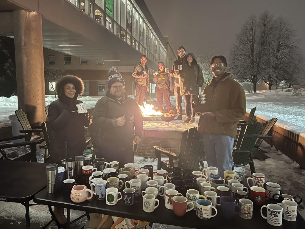 Snowy Background with a fire pit in the middle surrounded by 4 people with coffee mugs in hand, three more individuals in front of the fire fit with a table filled with coffee mugs