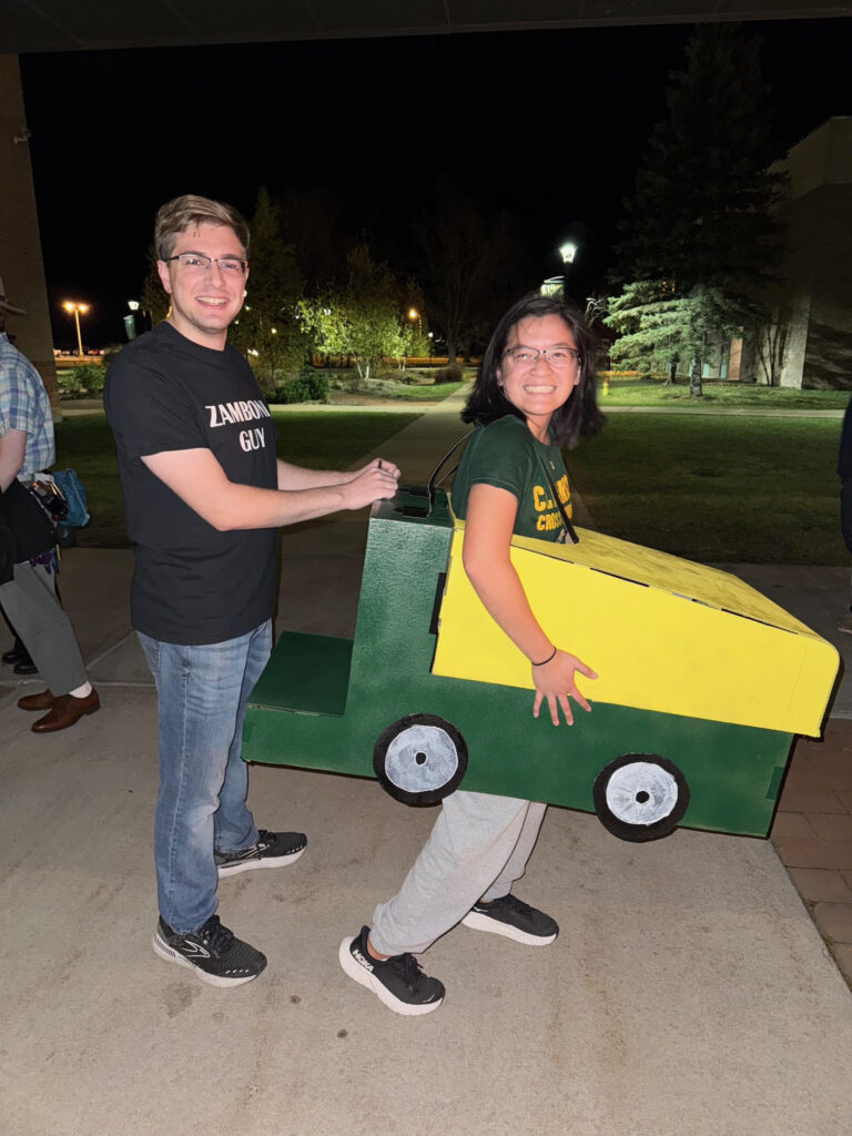 Two students dressed as a humorous duo for Halloween: one wearing a shirt labeled 'Zamboni Guy' stands behind another student who is wearing a green and yellow cardboard costume shaped like a Zamboni. Both are smiling and posing outdoors at night, with a campus pathway and trees visible in the background.